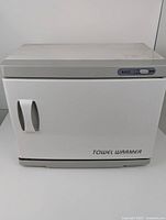 Closed towel warmer showing white rectangular body with dual curved handles on front door and power control switch on top right.