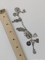 Sterling silver charm bracelet laid next to a ruler showing length slightly over 6 inches. Various detailed charms visible hanging from chain.