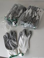 Photo showing two packs of white and grey work gloves with green elastic wristbands; one pack sealed, the other opened with loose gloves shown