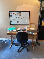 Wooden art desk with black rolling chair, standing lamp to the right, a large magnetic whiteboard mounted on the wall behind the desk with assorted magnets.