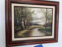 Framed landscape painting of wooded area and river hanging on wall