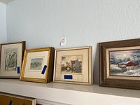 Photo showing six framed watercolor paintings resting on a white shelf, depicting various scenic village and winter landscapes.