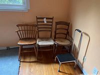 Four chairs with different designs on a wooden floor near a beige wall, including ladder-back and spindle-back styles plus a small black seat step stool with metal frame.