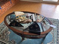 Full view of oval glass top wooden coffee table with decorative Asian fans placed on top and an area rug beneath