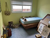 Bedroom corner showing twin mattress on brown frame with 4 hats, window with blinds, framed artwork on floor, and bags on carpet.