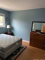 Full view of the bed with mattress and part of the wooden chest of drawers with mirror.
