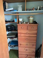 View of natural wood dresser with seven drawers in closet, various small items on shelves above and around it.