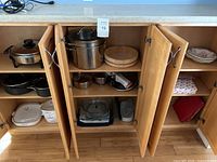 Three open wooden cabinets showing various cookware including Corning Ware ceramic dishes, stainless steel pots, and a wooden basket.