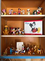 Full view of three shelves with various Disney character figurines and ceramic Mickey Mouse bank box.