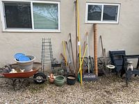 Full view of garden items arranged outside along a wall, showing wheelbarrow, planters, tools, plant supports, and other garden accessories.