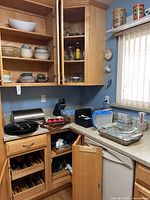 Wide angle showing kitchen cabinets with shelves holding ceramic and glass bowls, glassware, some condiments and other kitchen essentials, and countertop with knife block, glass dishes, storage containers.