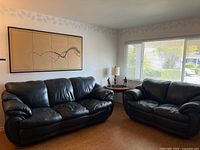 Two dark navy blue leather couches positioned in a corner of a room with a large window and framed wall art above the couch.