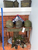 Three military helmets on wooden table with a folded American flag in display case behind. Various military gear items on brick hearth below.