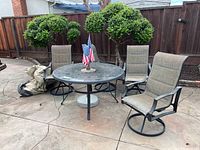 Patio furniture set with glass table and four swivel chairs shown outdoors on a concrete patio with wooden fence and greenery in the background.