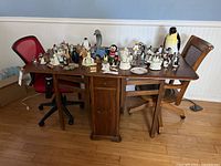 Photo of wooden drop-leaf table with various figurines on top and two mismatched chairs, one wooden and one office chair.