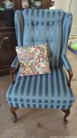 Front view of the turquoise striped 1960s wing back armchair with floral throw cushion on the seat, showing exposed wood on arms and legs and upholstery.