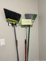 Five cleaning tools including three brooms, a mop/squeegee, and a small hand broom hanging on a wall rack.