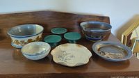 All seven pottery pieces displayed on wooden surface: bowls, plates, pedestal dishes with varied glazes and patterns.