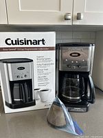 Full shot of Cuisinart DCC-1200C 12-cup programmable coffeemaker next to its original box. A reusable gold-tone coffee filter and charcoal water filter are positioned in front of the machine.
