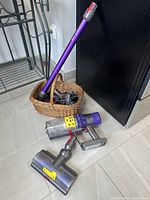 Dyson vacuum parts arranged on floor including main vacuum body, motorized brush head, purple wand, and wicker basket with accessories and charger