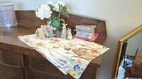Table runner with bunny and floral embroidery laid out on wood dresser with various bunny figures and artificial flowers in a lettuce pitcher