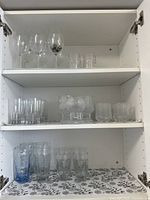 Open cabinet shelf showing upper and middle shelves with various clear glasses including stemmed wine glasses and patterned drinking glasses.