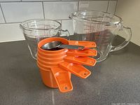 Photo of 2 Pyrex glass measuring cups (2 cup and 4 cup) with 5 stacked vintage orange Tupperware measuring cups and stainless steel 1/8 cup measuring spoon on countertop.