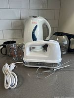 Lot showing Cuisinart hand mixer, Sunbeam electric kettle, glass coffee pot, Fire King measuring cup, and stainless steel sifter on kitchen counter.