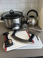 Photo showing entire kitchen lot including Crock-Pot slow cooker, kettle, chopping cradle, cutting utensil sharpener, stainless steel scraper, two cutting boards, and fridge thermometer.