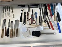 Top-down photo of drawer showing varied kitchen utensils arranged neatly in white drawer organizer. Includes spreaders, kitchen scissors, small whisks, bottle openers, and spatulas.