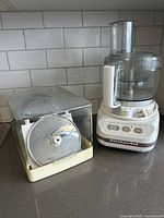 KitchenAid Ultra Power food processor with clear plastic bowl and feeding tube next to an organizing case holding multiple slicing/shredding discs.
