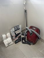 Overview shot showing red canister vacuum, grey hose, telescopic wand, assorted attachments, and sealed Miele bag packs