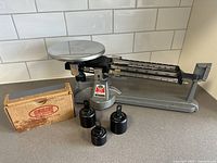 Full view showing Ohaus triple beam scale with round platform and three calibration weights including original box.