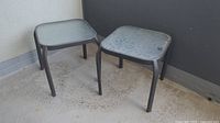 Photo showing two square glass top patio side tables with black metal frames sitting on a concrete balcony floor, dusty and in need of cleaning.