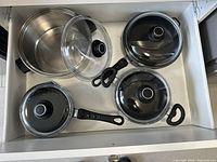 Drawer with 4 pots visible: 3 black cast iron pots with glass lids and one stainless steel pot without lid, plus an extra lid and pot handle inserts.