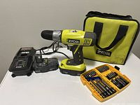 Full lot showing Ryobi cordless drill, batteries, charger, Dewalt bit set, and Ryobi carrying case on a white surface.