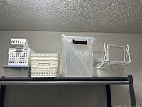 Storage containers and kitchen cupboard shelves displayed on a shelf: clear plastic bin, white plastic baskets, beige woven basket, and metal wire shelves.
