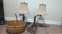 Pair of metal base table lamps with beige lampshades and two baskets placed on floor