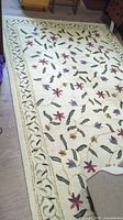 Full view of cream colored wool area rug with floral vine pattern laid on floor beside furniture.