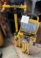 Two yellow adjustable height work light stands, one with a light fixture cage and power cord, one without light fixture, folded and placed next to each other on floor.