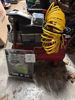 Red air compressor unit with attached yellow coiled hose and pressure release tube visible; Grip Air Tank Assembly Kit package visible in front