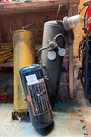 Vertical gray air tank with two pressure gauges and fittings next to vertical yellow air tank; blue horizontal air tank in front on floor