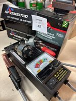 Two black boxes labeled NP Boosted with ignition coils, a battery tester device with analog gauge and connected clamps, and a 3M fiberglass repair kit box.