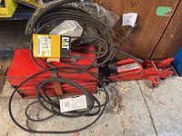 Multiple various sized belts mostly CAT brand wrapped and loose on and beside a red hydraulic trolley jack missing pole.