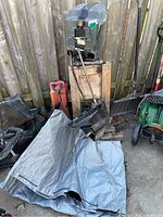 Outboard motor mounted on wooden stand with attached lower unit, orange gas can and grey protective cover partly covering parts of the lot, set beside wooden fence outdoors.