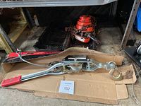 1 ton cable puller with red handles laid on cardboard with chain hoist visible in the background.