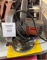 Front and side view of Black & Decker circular saw showing the yellow toothed blade and power cord in the foreground, with sanding belts in the background.
