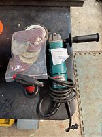Makita disc grinder with attached power cord and black handle, torch lighter, and plastic bag containing sanding discs.