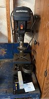 Full view of Mastercraft drill press showing base, column, motor housing with Mastercraft logo, and two lever handles for operation. Drill press is placed on a blue workbench surface.