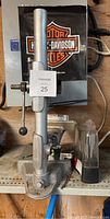 Metal Sears drill press with lever, base, and clamp, shown on a shelf with a Harley Davidson bag in background.